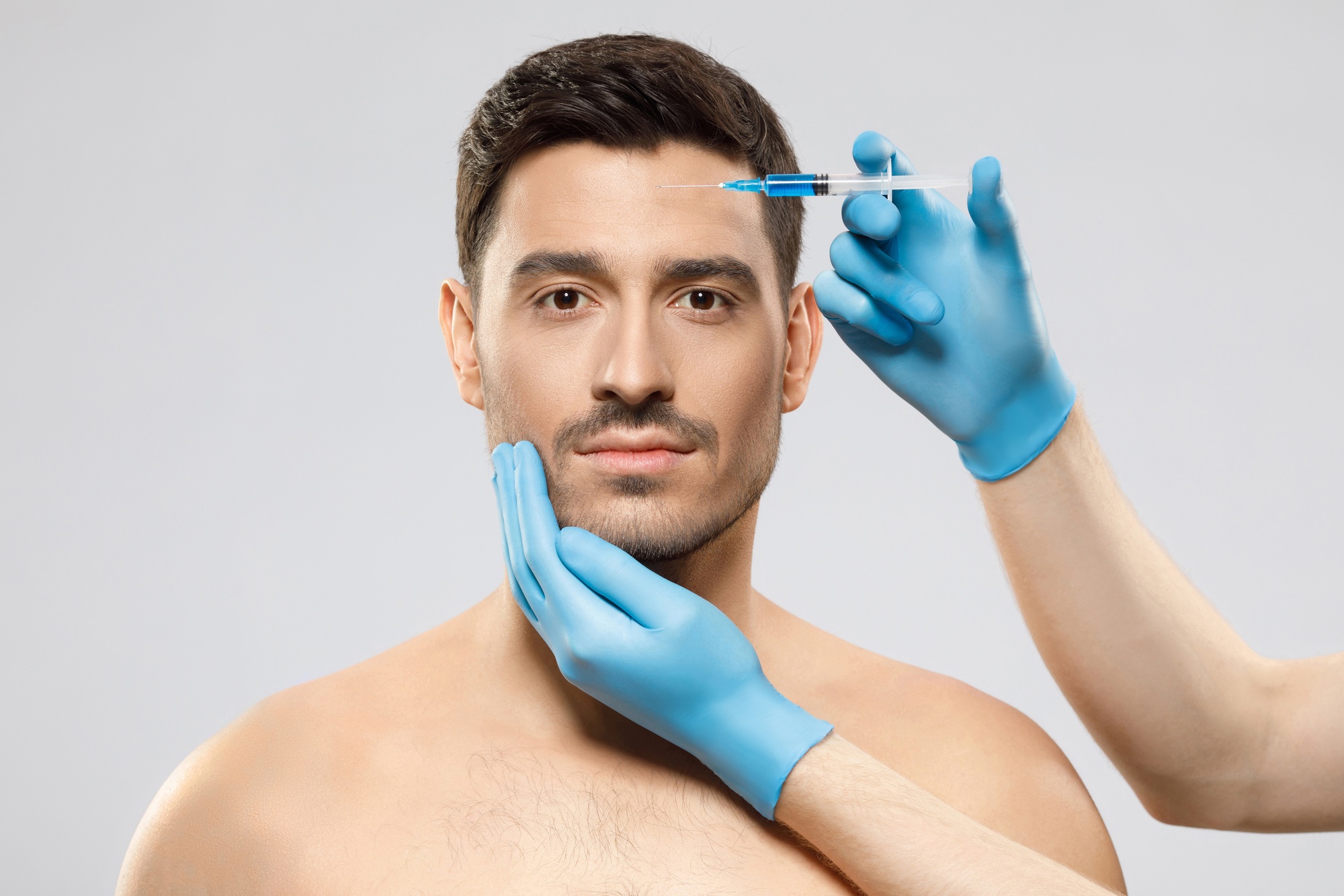 Young man isolated on gray background looking at camera while receiving botox into forehead